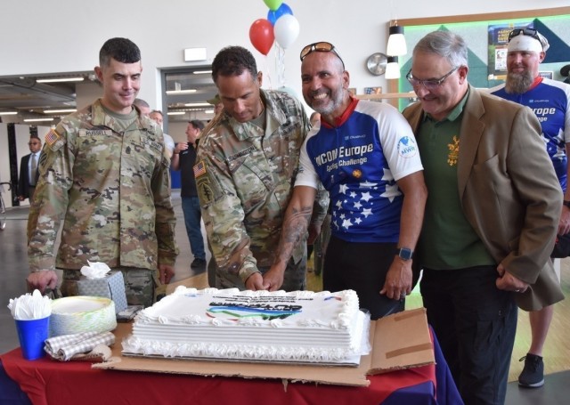 Wiesbaden Garrison Command Sergeant Major Richard Russell, left, and Steven Steininger, right, a volunteer sport shooting instructor, watches as Garrison Commander, Col. Mario Washington, and Luis Castro, a member of the cycling challenge team, cuts the cake at the MWR Outdoor Recreation's grand opening Sept. 7, 2021.